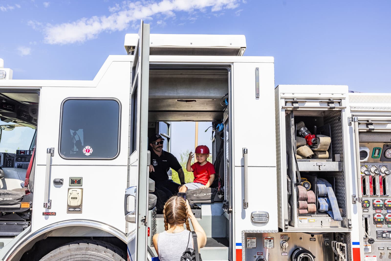 A young child in a red firefighter helmet sits in the cab of a Phoenix fire engine while a firefighter looks on.