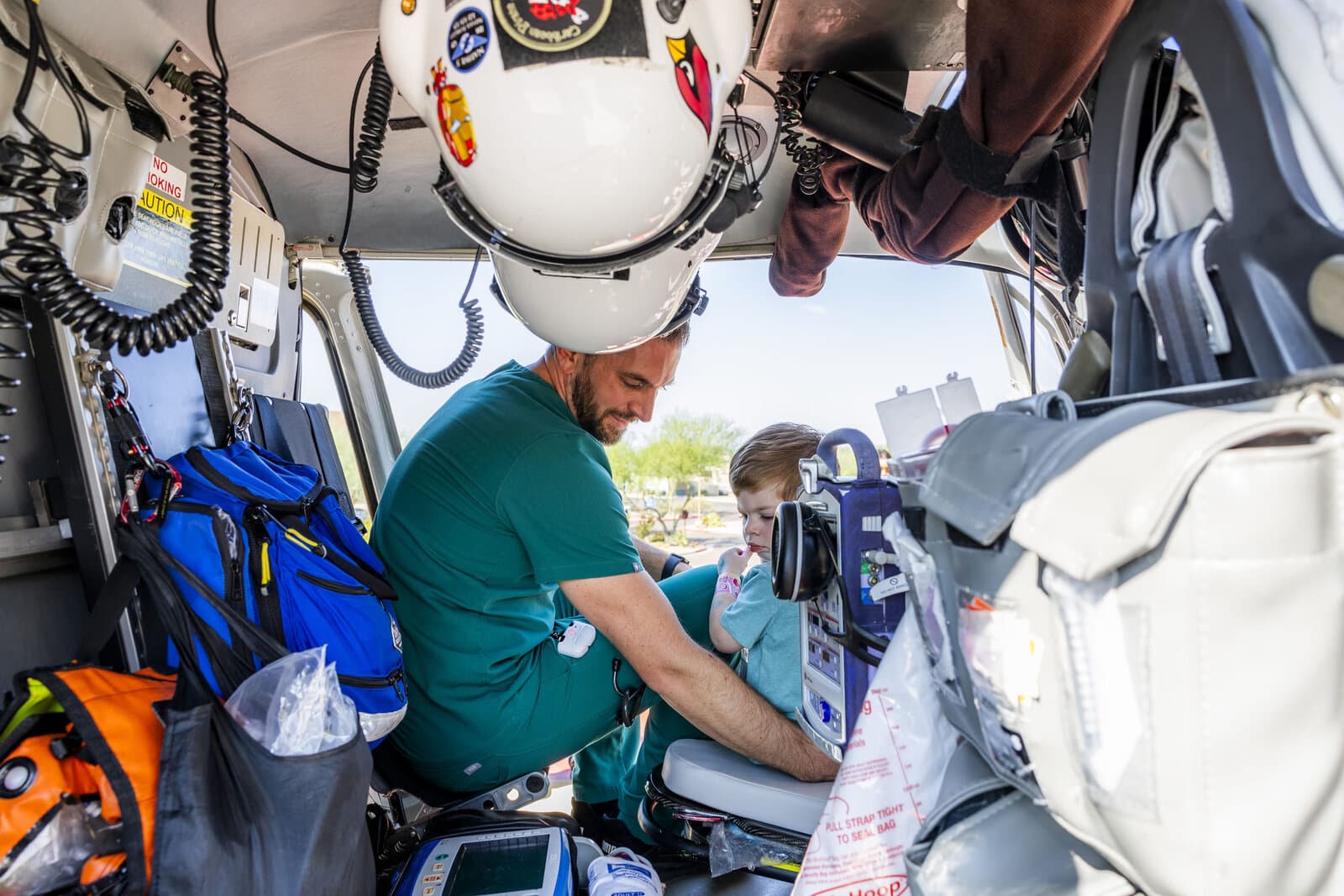 A flight medicine physician in scrubs and a pilot helmet helps a toddler buckle into the cockpit of a medical helicopter.