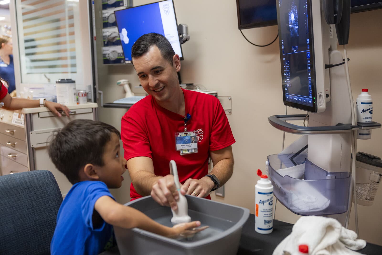 A physician guides a child's hand on an ultrasound probe with the live image displayed on a monitor behind them.