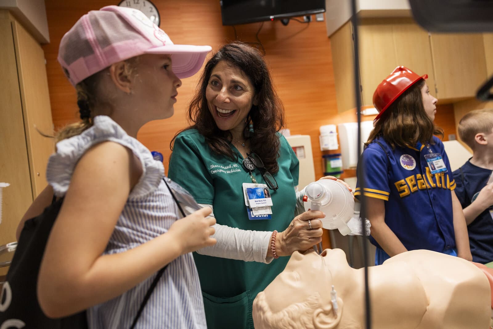 An emergency physician in scrubs shows a child how to bag-mask ventilate a mannequin while other kids in fire helmets and uniforms look on.