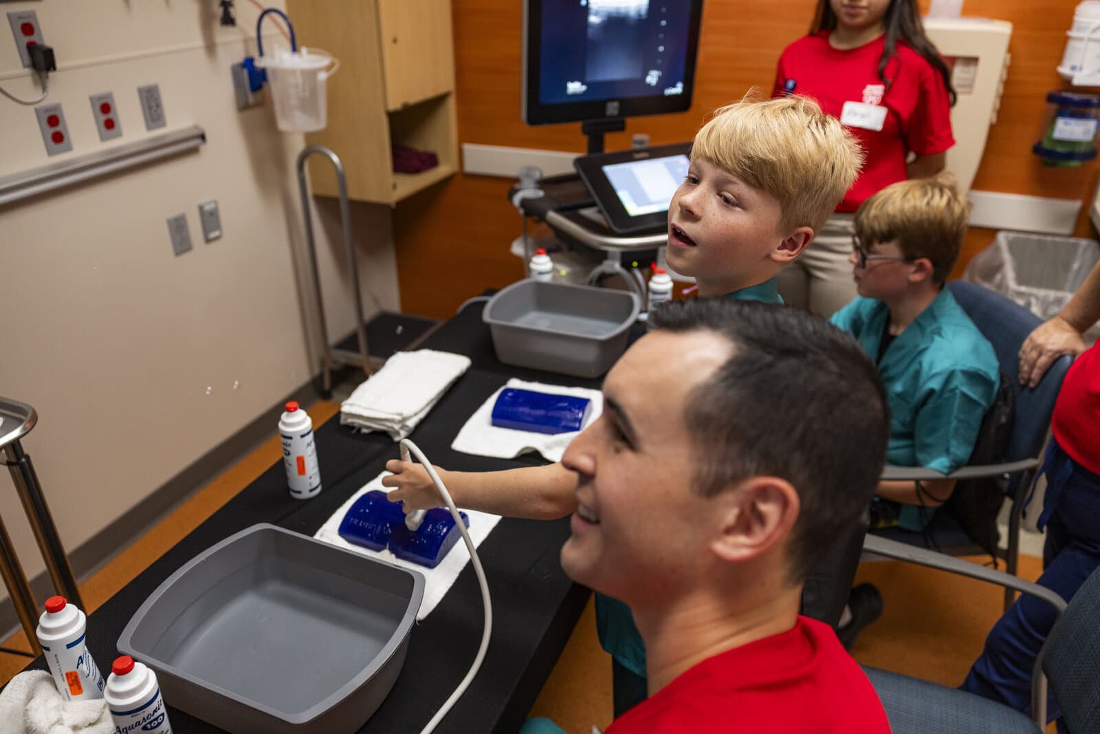 A boy holds an ultrasound probe to a training phantom while another physician watches the screen.
